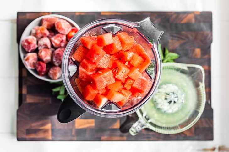 Watermelon cubes in the blender with lemon juice and frozen strawberries sitting next to it on a wooden cutting board.