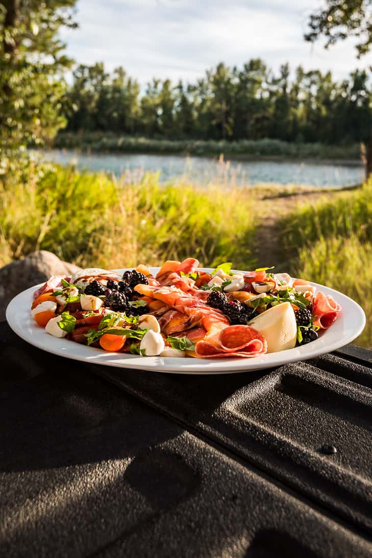 An oval platter of Summer Antipasto sitting on the corner of a truck tailgate with a river and trees in the background.