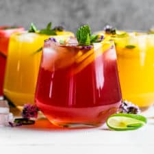 Three red and two orange colored mocktails in cocktail glasses on a white background.