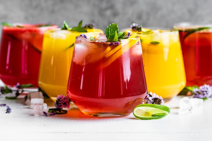 Three red and two orange colored mocktails in cocktail glasses on a white background.