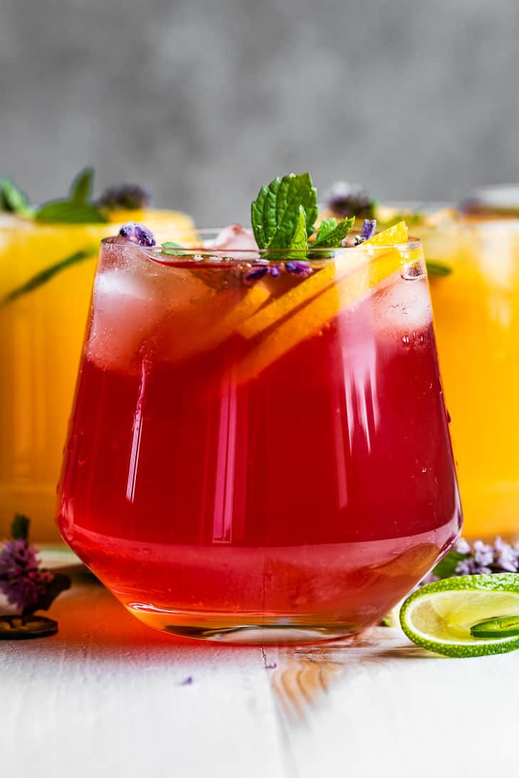 Red and orange colored Kombucha Mocktails in cocktail glasses topped with mint and orange slices on a white background.