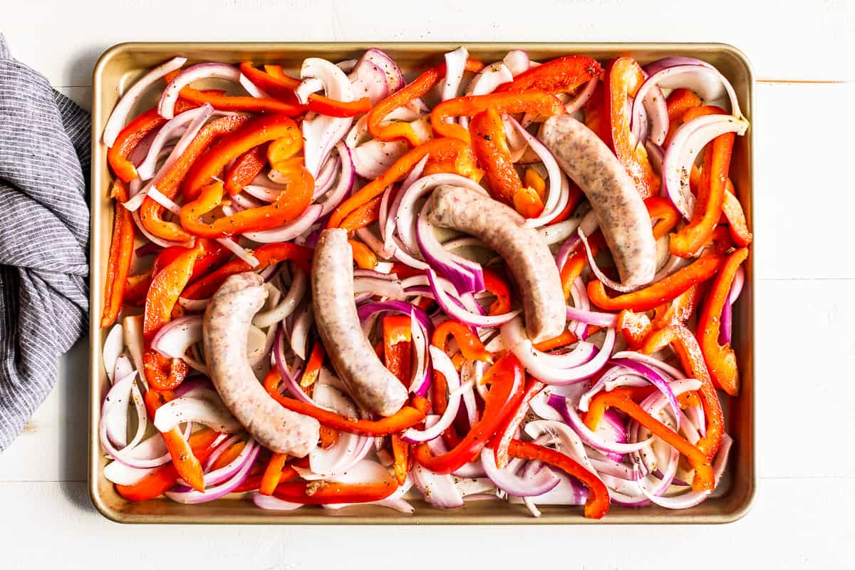 Peppers and onions on a cookie sheet with sausages on top before baking.
