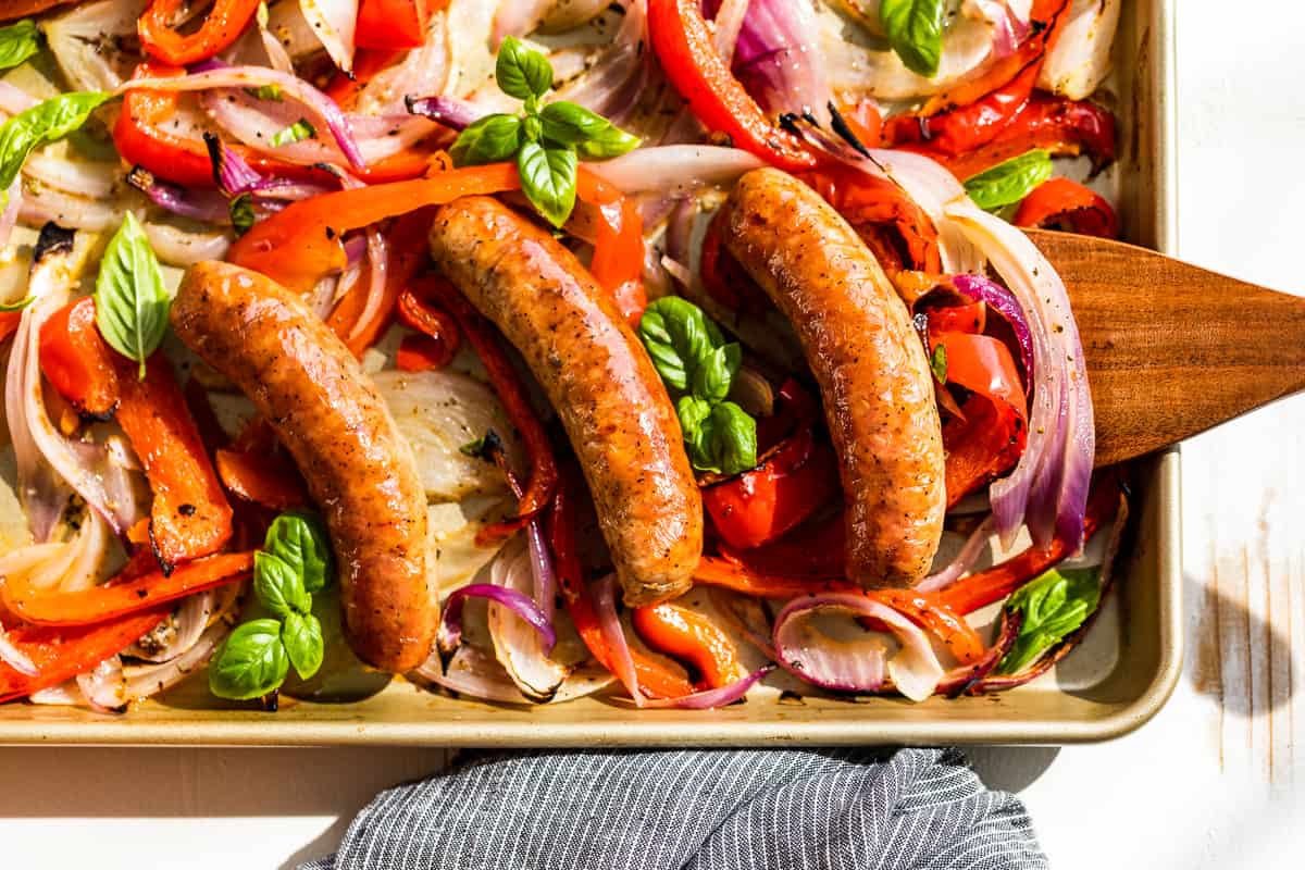 Sausage and Peppers on a gold sheet pan garnished with sprigs of basil.