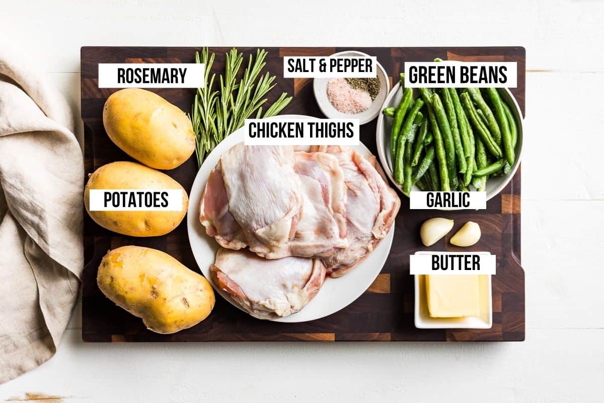 Chicken thighs, potatoes, green beans, garlic, butter, and rosemary on a wooden cutting board.