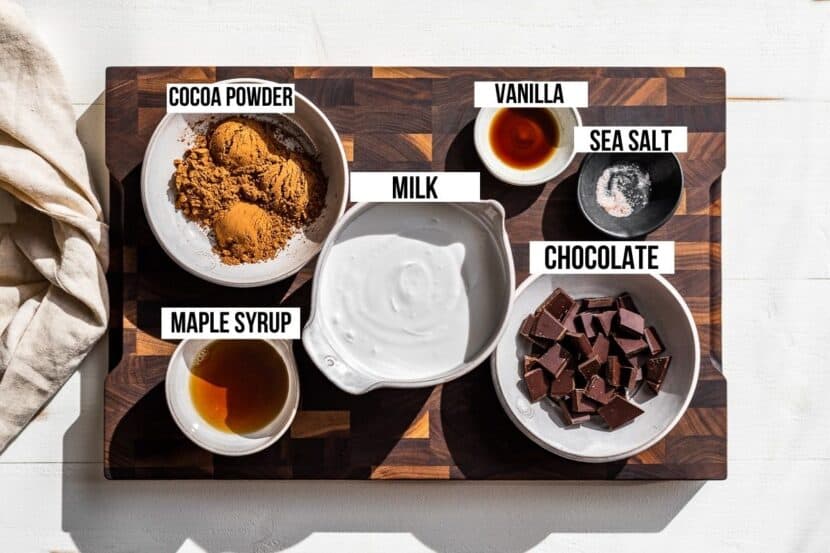 Cocoa powder, maple syrup, milk, vanilla, and chocolate on a wooden cutting board.