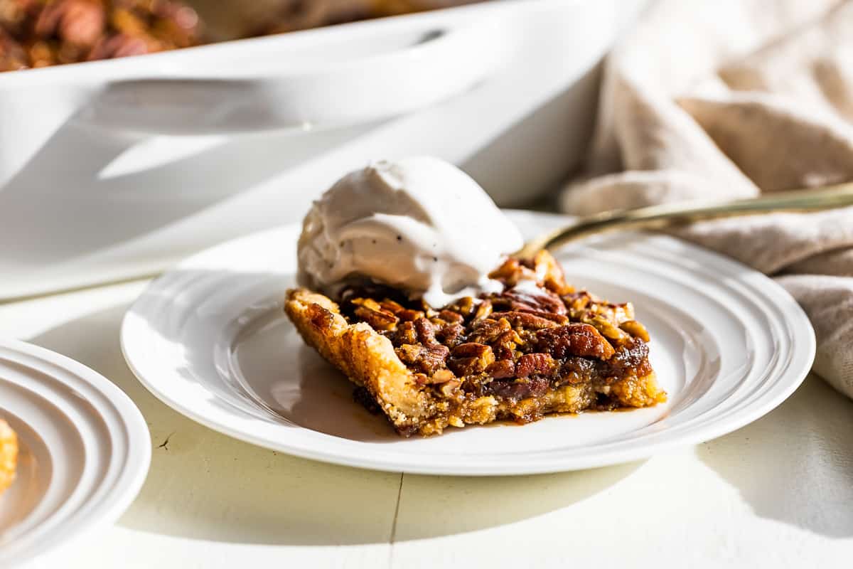A pecan pie bar on a white plate with a scoop of vanilla ice cream on top.