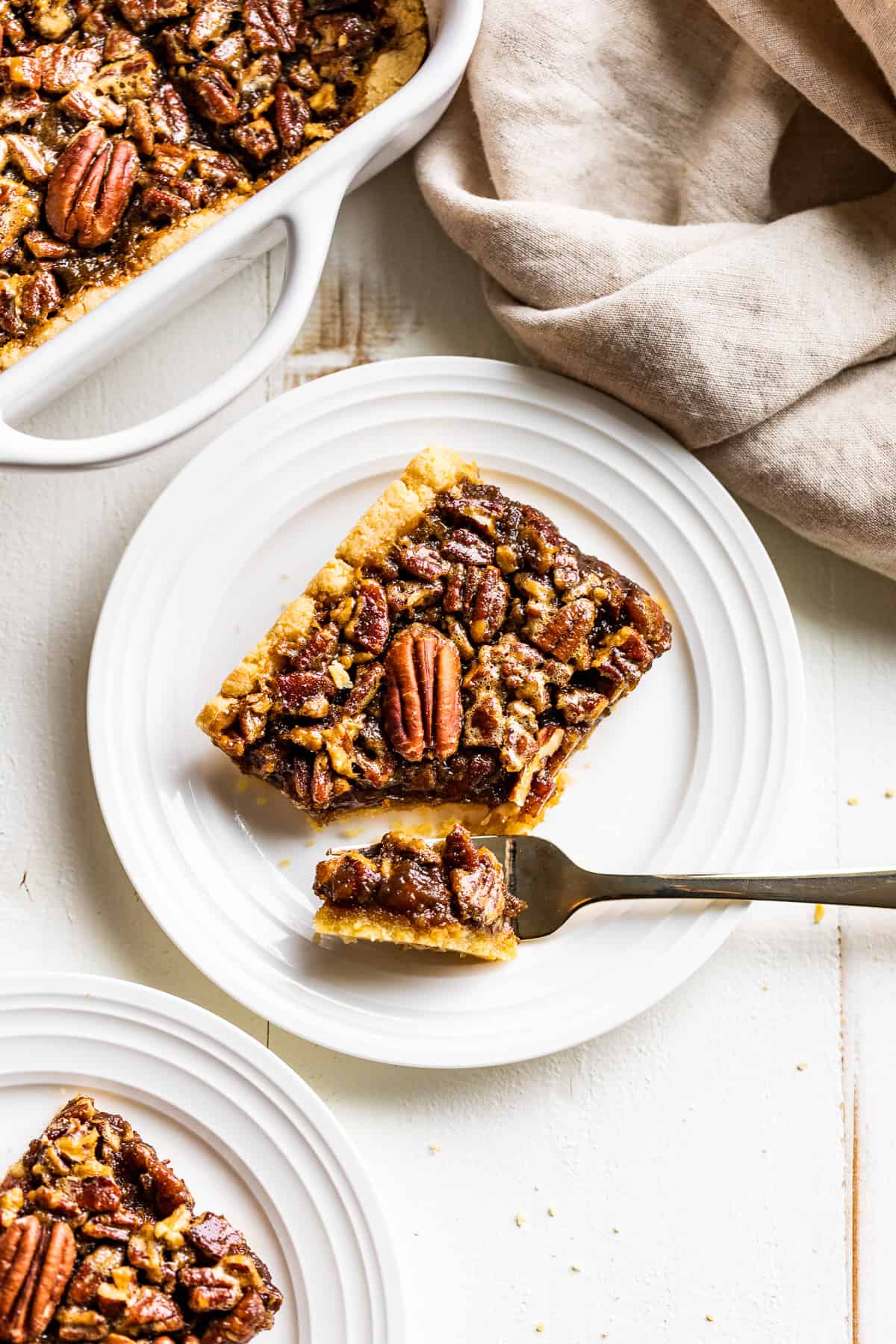 Two plates of pecan pie bars next to the pan of pecan pie bars with a tan linen on the side.