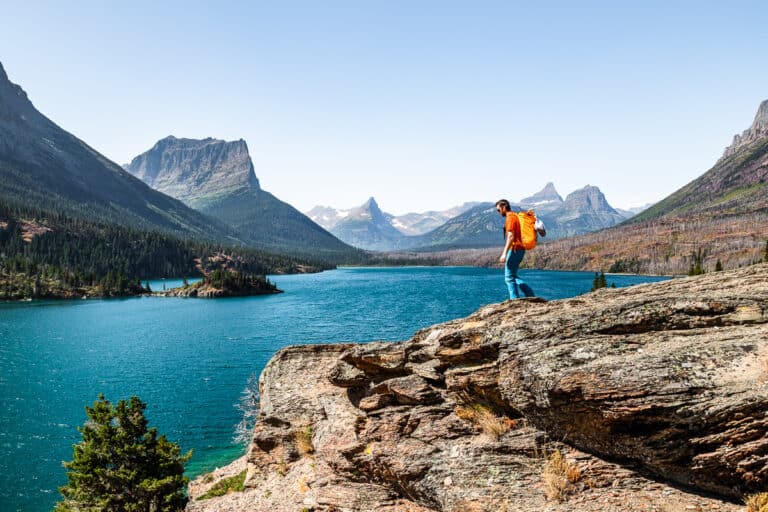 A man with an orange backpack walking down a rocky slab overlooking a lake and mountains.