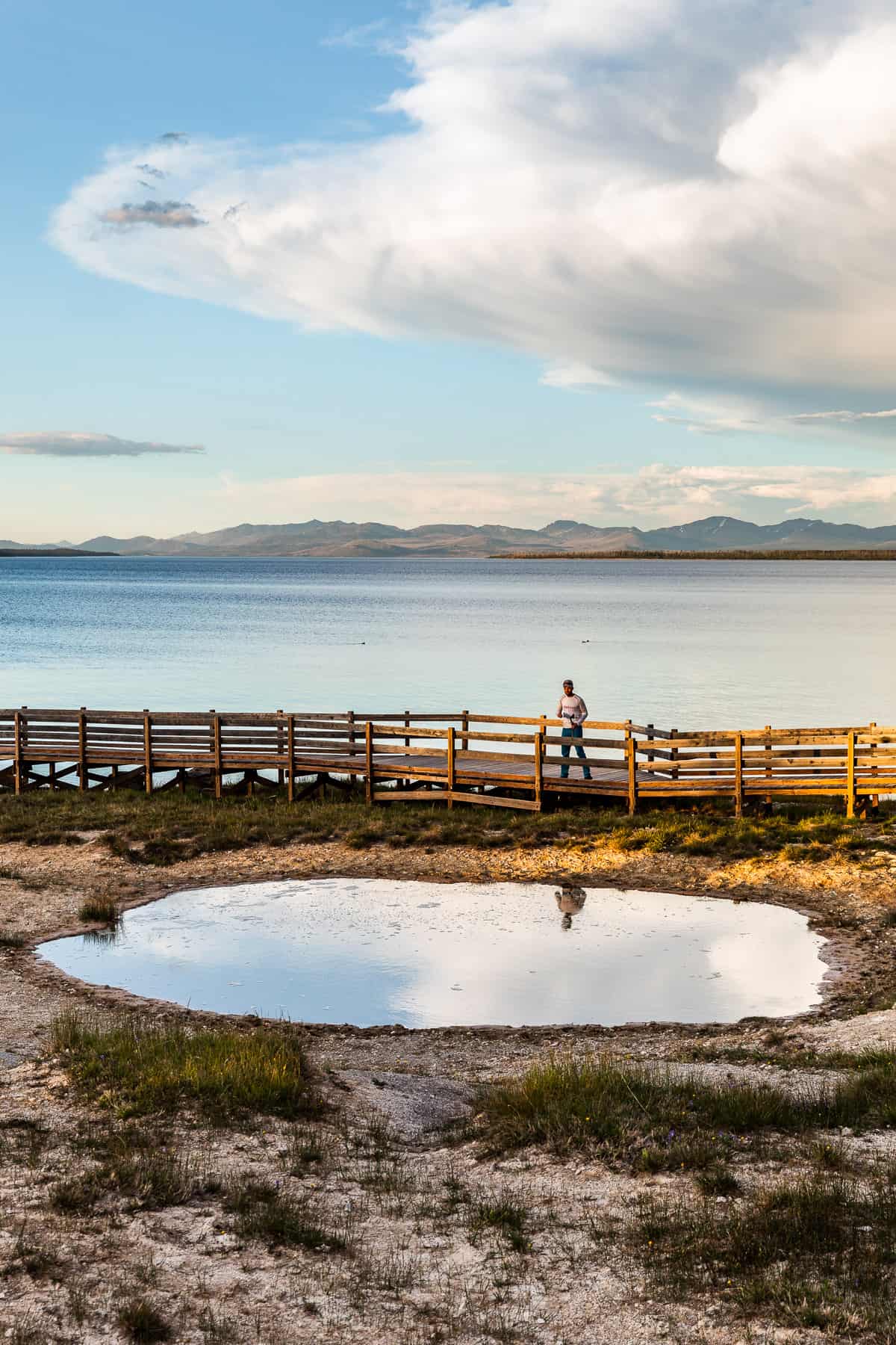 A man leaning on the railing of the boardwalk looking down into a geyser with Yellowstone Lake in the background.