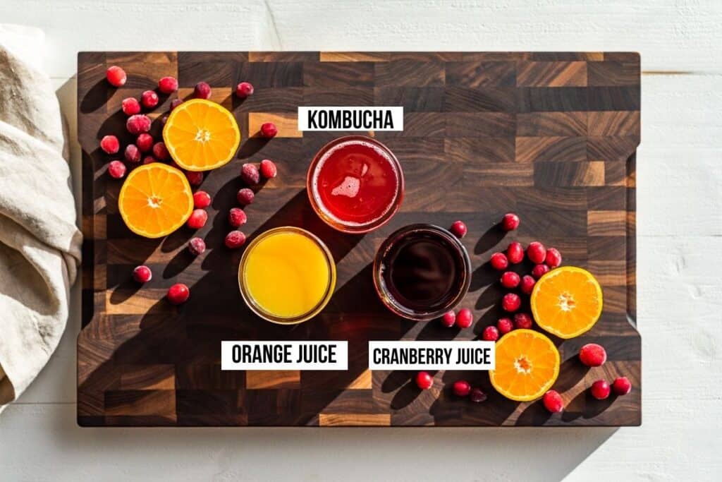 Kombucha, Orange Juice, and Cranberry Juice in jars on a wooden cutting board with orange slices and cranberries.