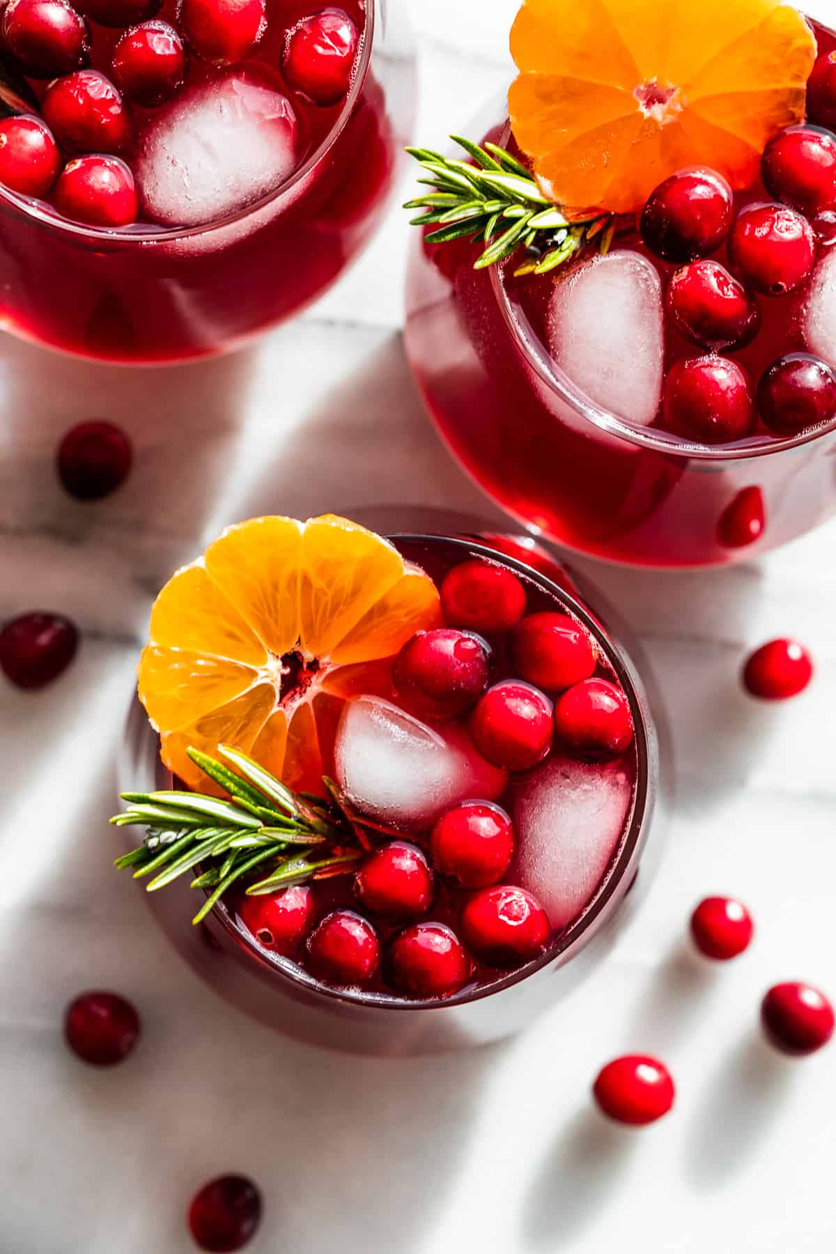 Straight down view of three glasses mocktails topped with cranberries, orange slices, and rosemary sprigs.