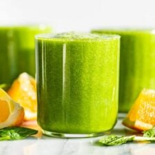 Three glasses of Detox Green Smoothie with orange wedges and spinach leaves around the glasses.