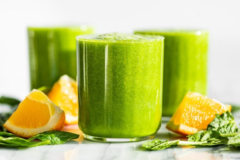 Three glasses of Detox Green Smoothie with orange wedges and spinach leaves around the glasses.