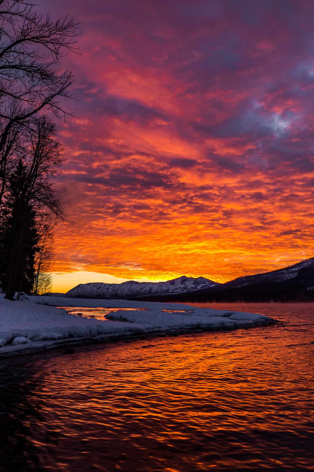 Fiery orange sunset with bright pink high in the sky with a snowy lake shoreline.
