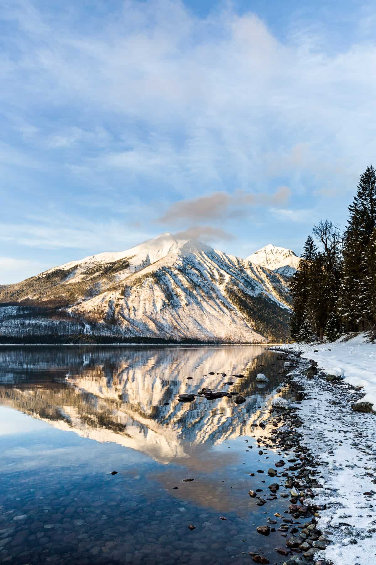 Mountain reflecting into a lake with blue skies and high white clouds and snow on the shoreline.