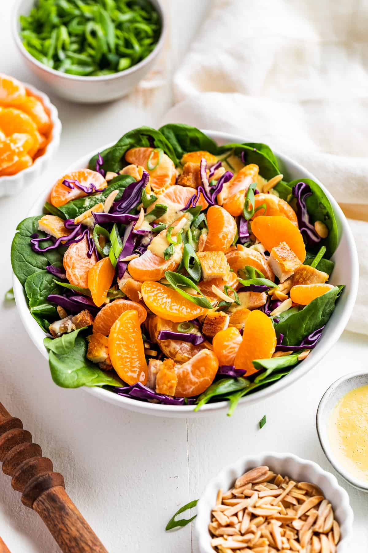 Side view of Mandarin Chicken Salad with wooden serving spoons, green onions, slivered almonds, and Mandarin oranges on the side.