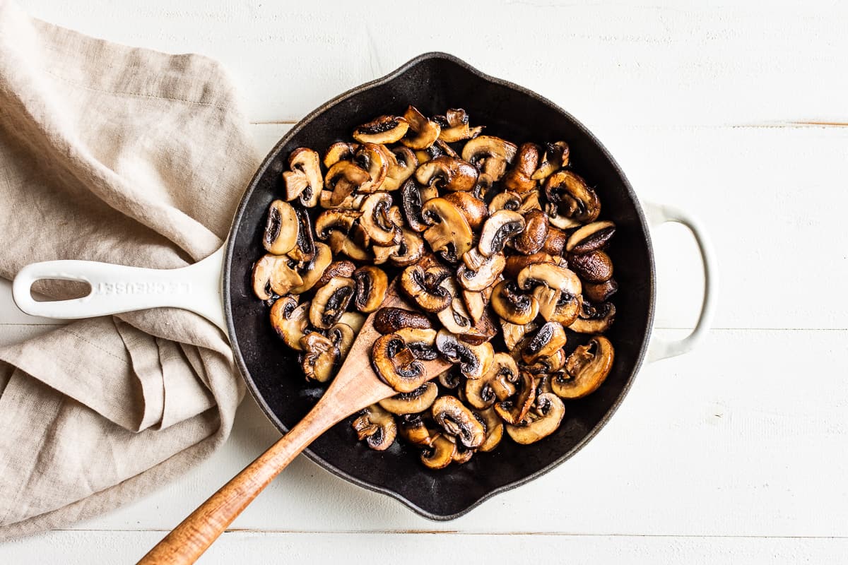 Sautéed mushrooms in a white skillet with a wooden spoon.