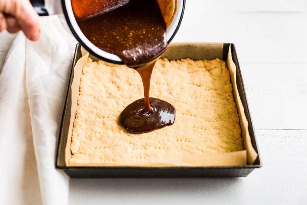 Caramel filling being poured over the baked and cooled shortbread crust.