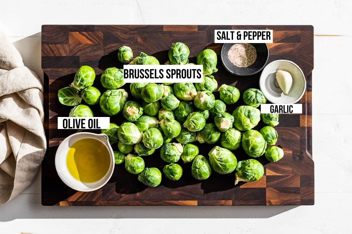Brussels sprouts, olive oil, garlic, sea salt and pepper in bowls on a wooden cutting board.