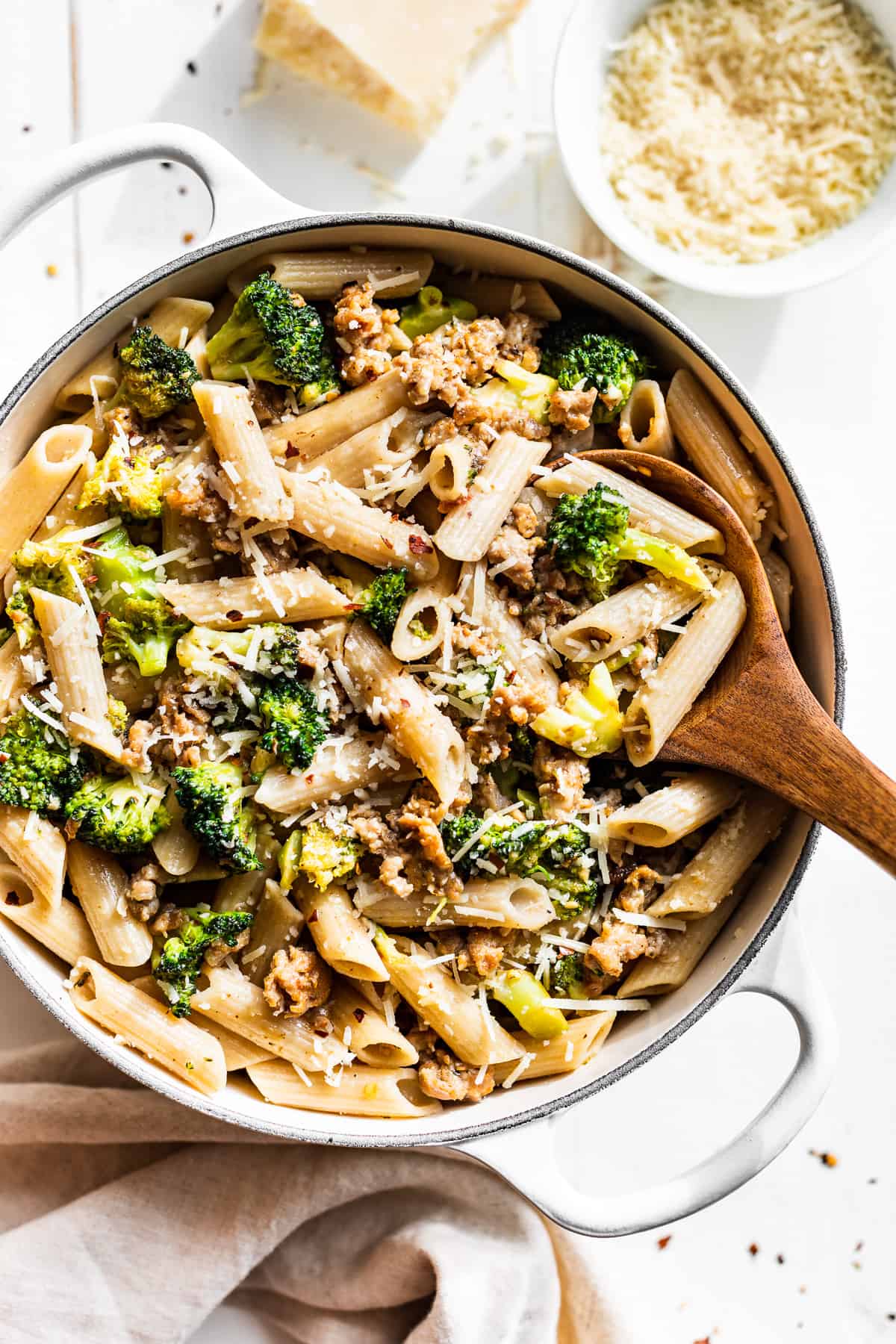 White pot filled with Sausage Broccoli Pasta with a wooden spoon and parmesan on the side.