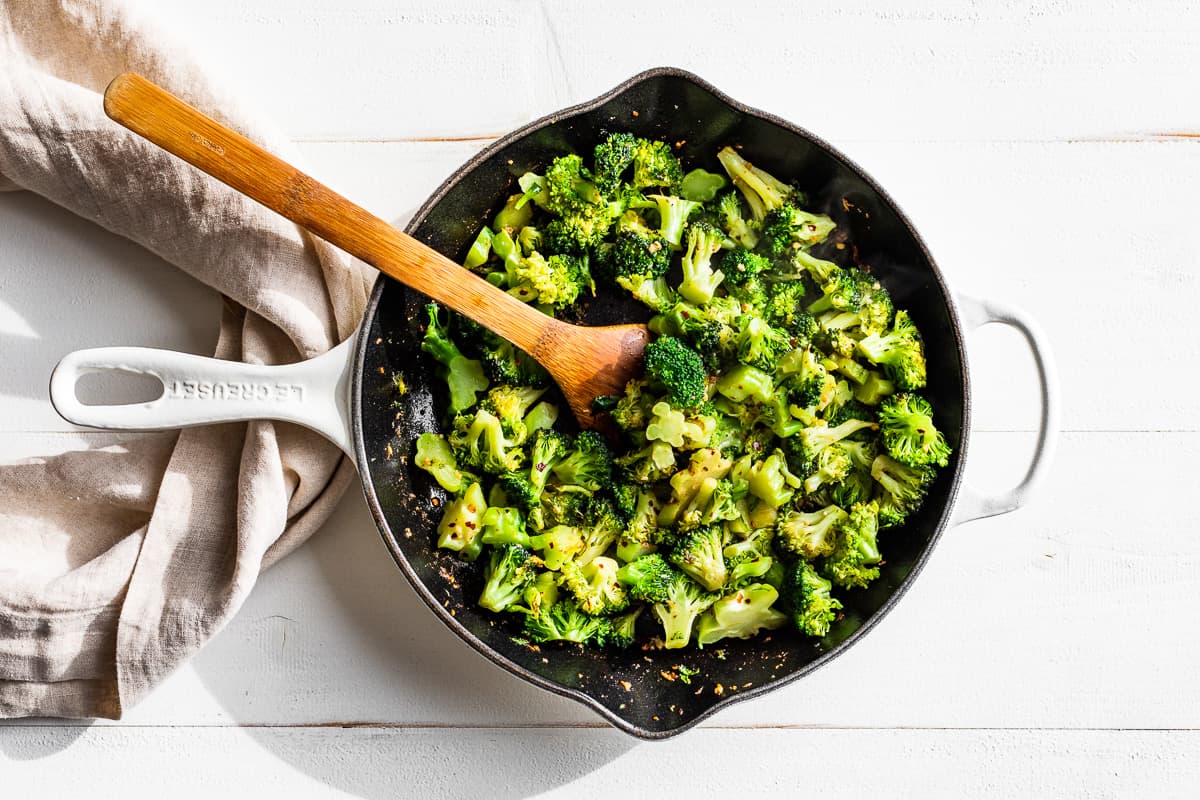 Cooked broccoli in a white cast iron skillet with a wooden spoon.