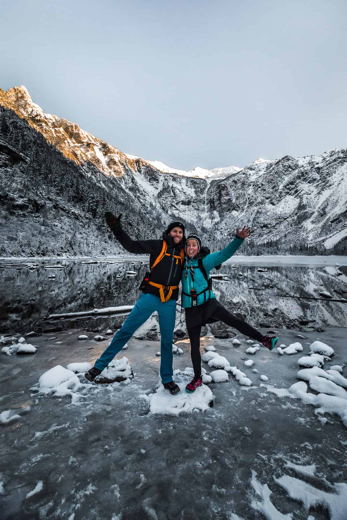 A snowy mountain cirque with an icy lake below and a couple standing at the edge of the lake.