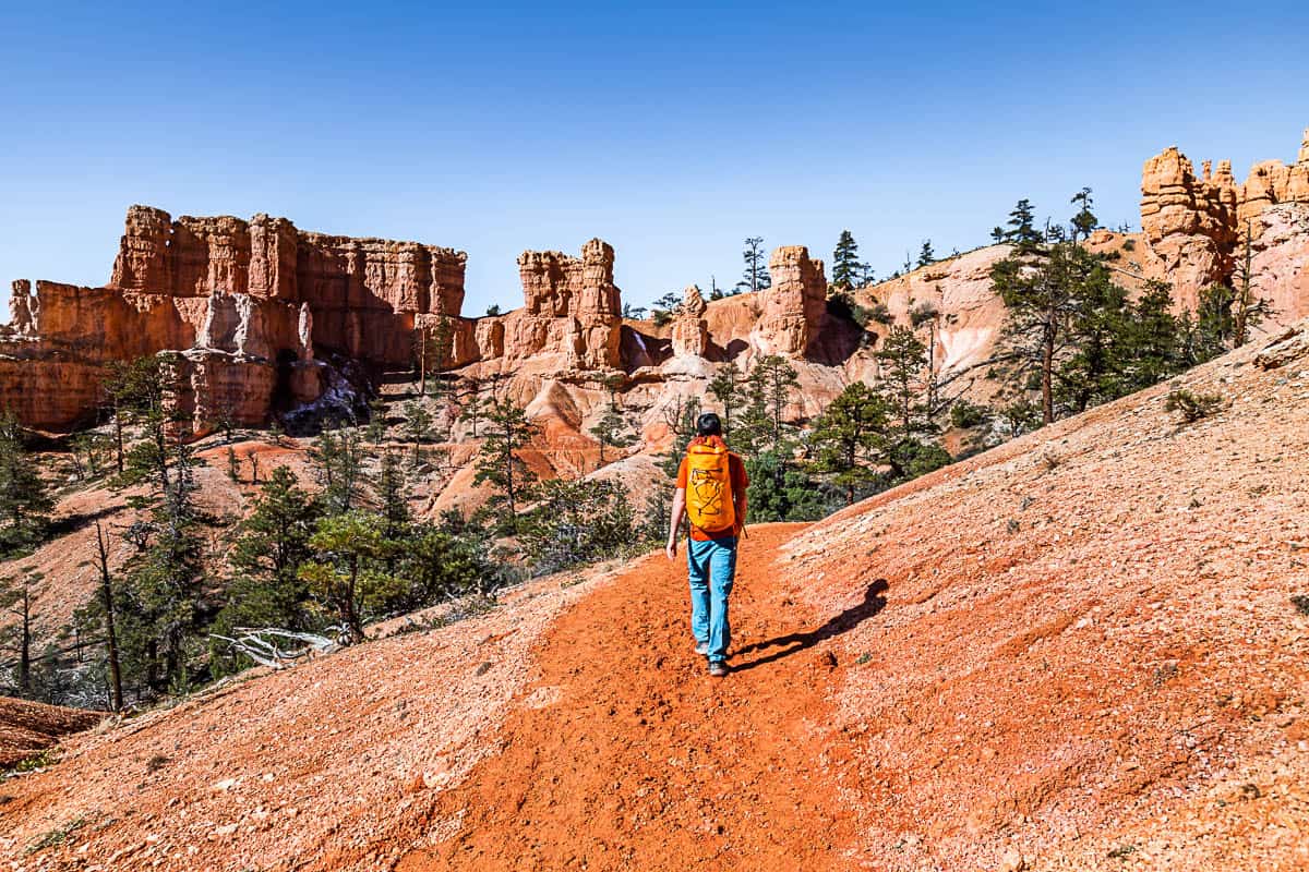 Man hiking with an orange backpack on an orange dirt trail with hoodoos in the distance and blue skies.