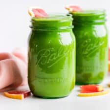 Two green glass jars of Grapefruit Green Smoothie with a blush linen napkin folded on the side.