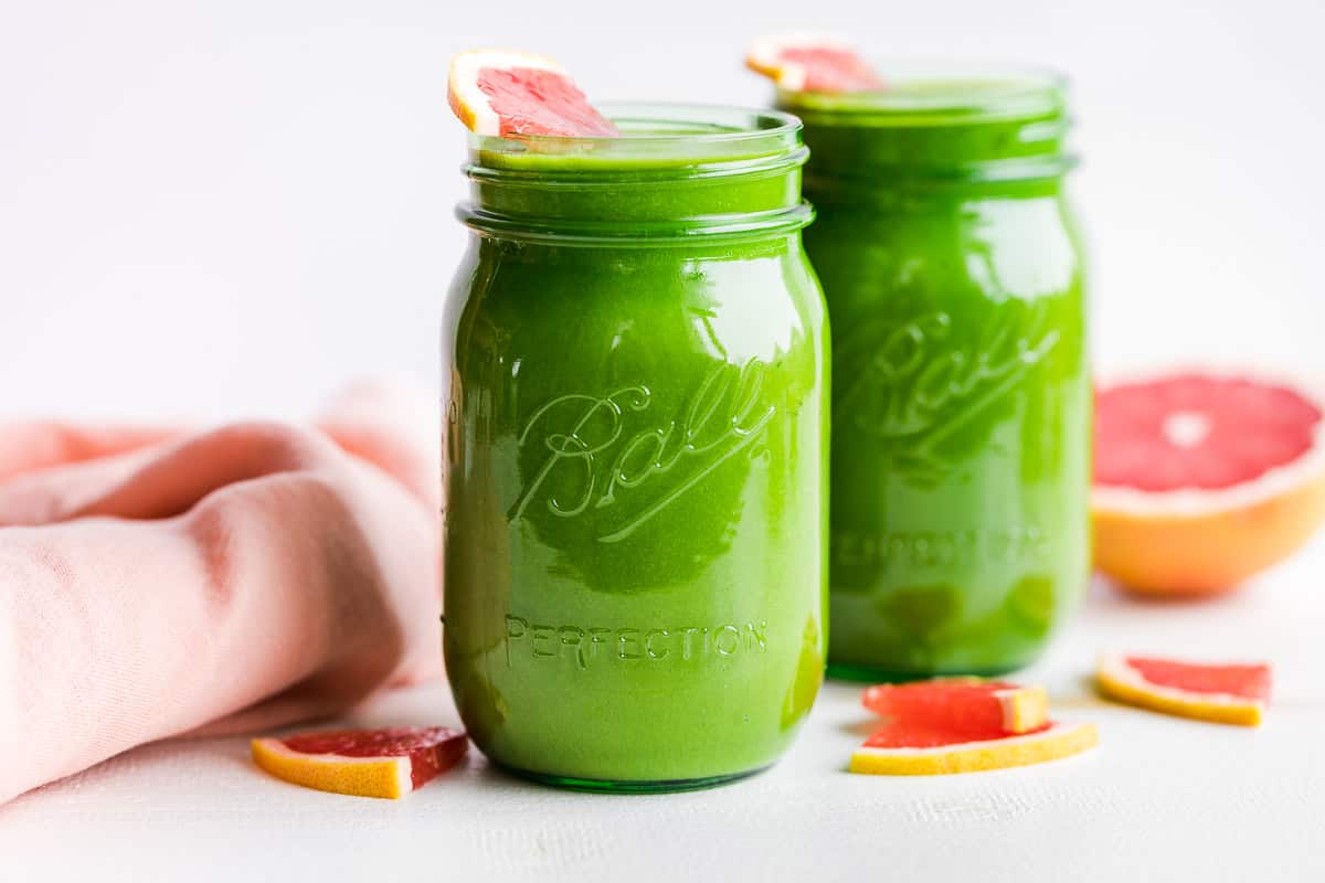 Two green glass jars of Grapefruit Green Smoothie with a blush linen napkin folded on the side.