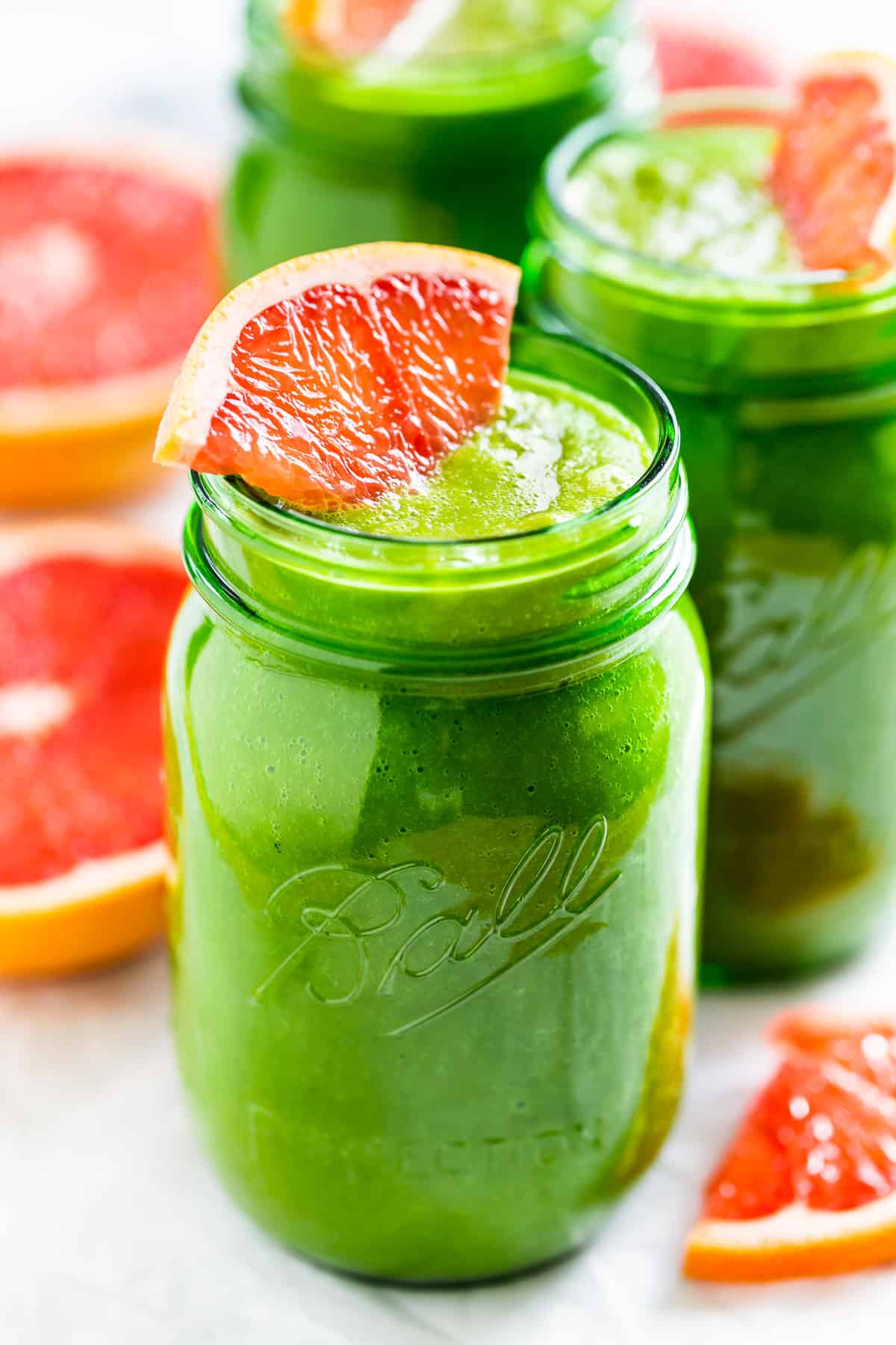 Three green glass jars of Grapefruit Green Smoothie with halved grapefruits on the side.