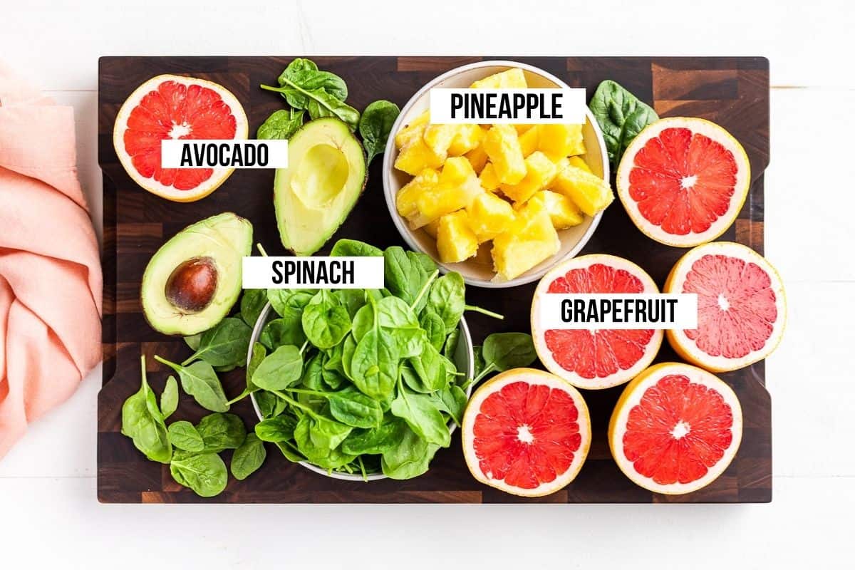 Halved grapefruits, frozen pineapple, avocado, and spinach on a wooden cutting board.