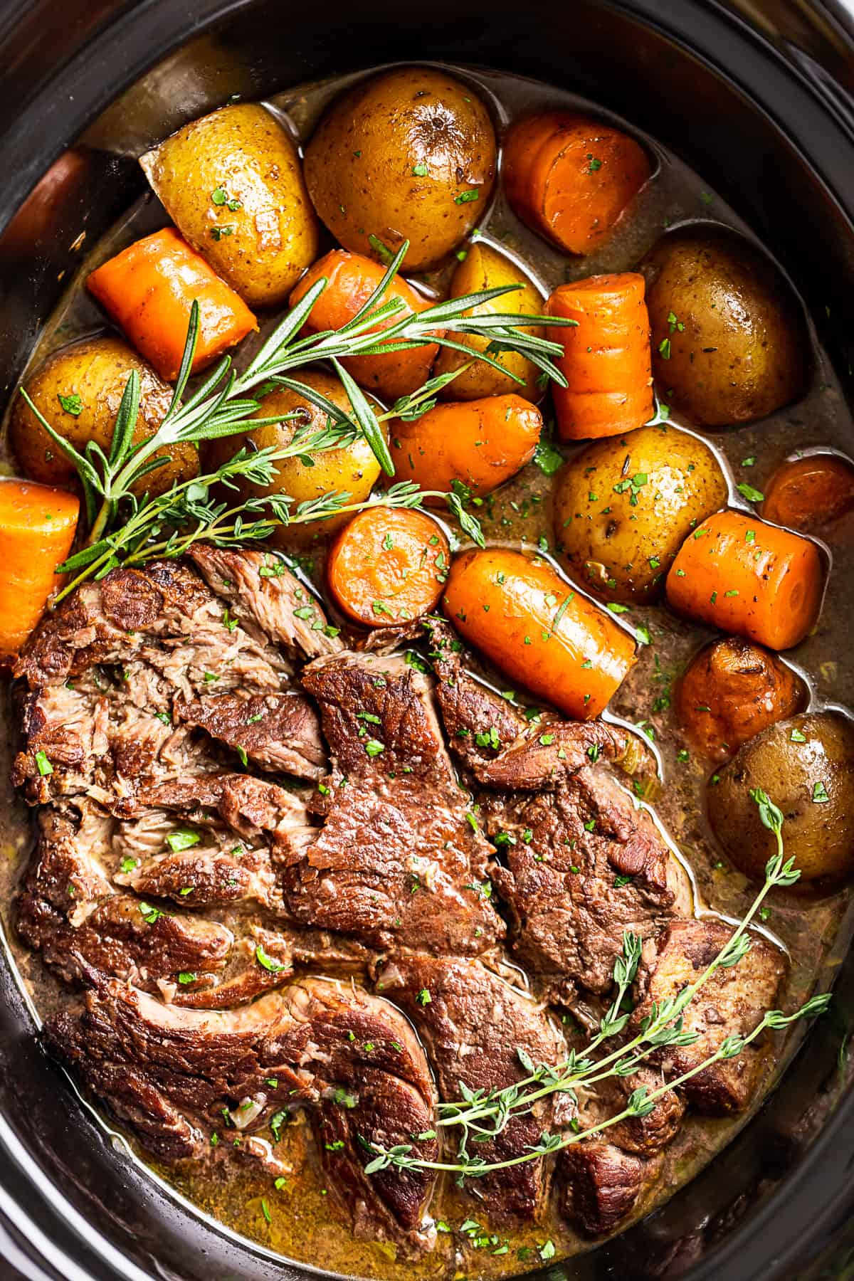 Close up view of Slow Cooker Pot Roast with potatoes, carrots, and herbs in a black slow cooker bowl.