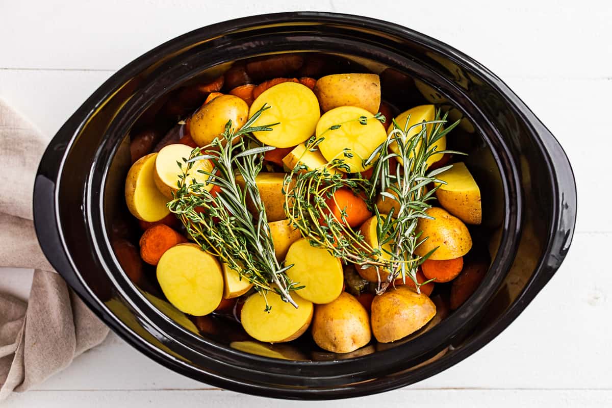 The beef roast in the slow cooker bowl topped with potatoes, carrots, and fresh herbs.