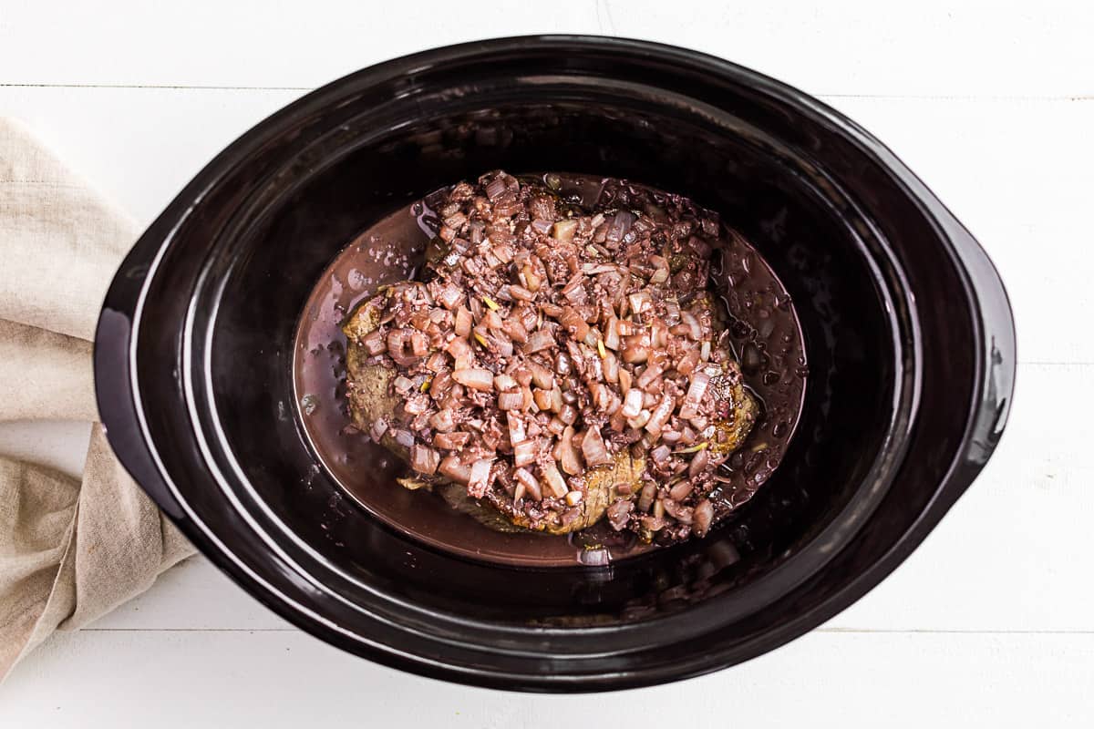 The beef roast in a slow cooker bowl topped with onions and garlic.