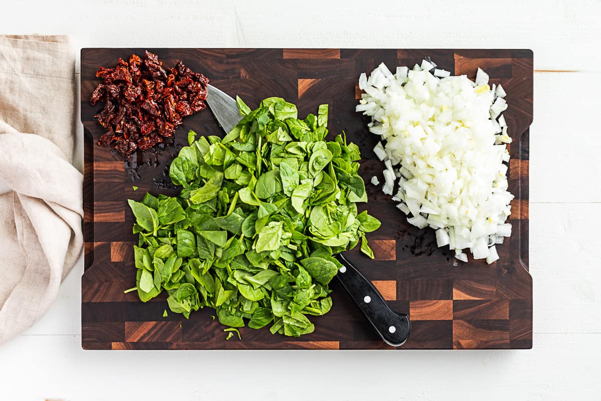 Chopped spinach, onions, and sun dried tomatoes on a wooden cutting board.