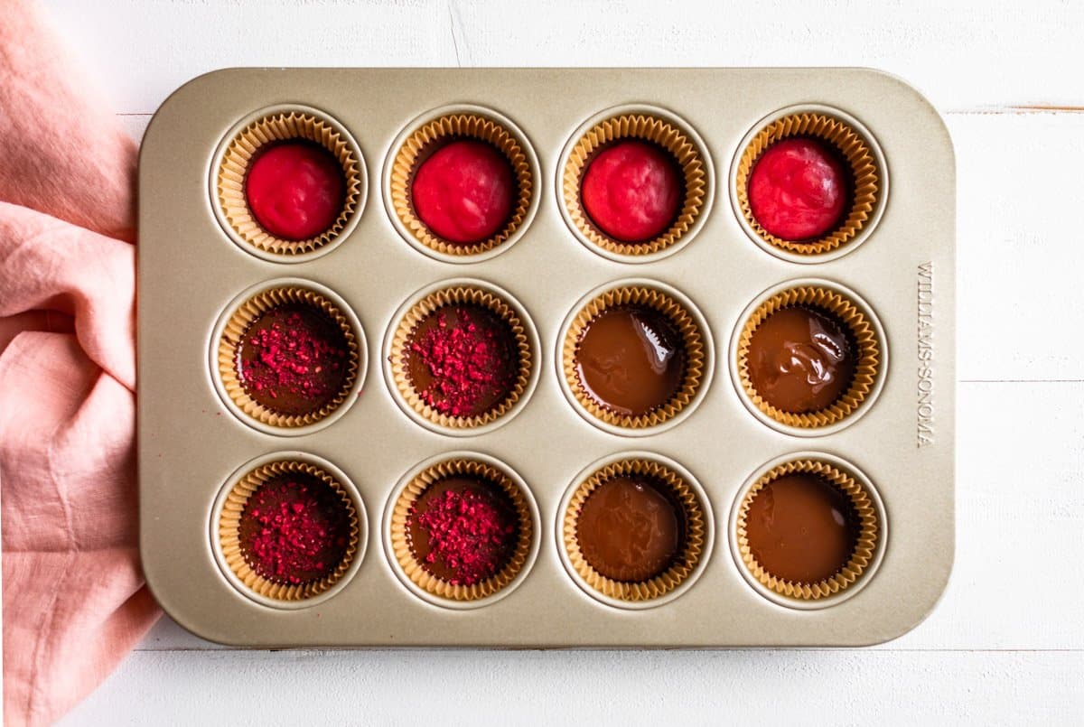 A muffin tin with muffin cup liners, some have melted chocolate, the others have the raspberry filling added.