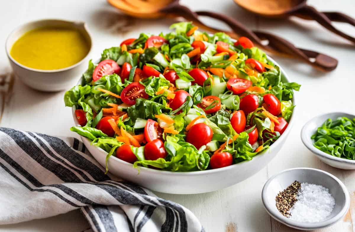 Finished salad in a white serving bowl with lemon vinaigrette on the side and wooden serving spoons.