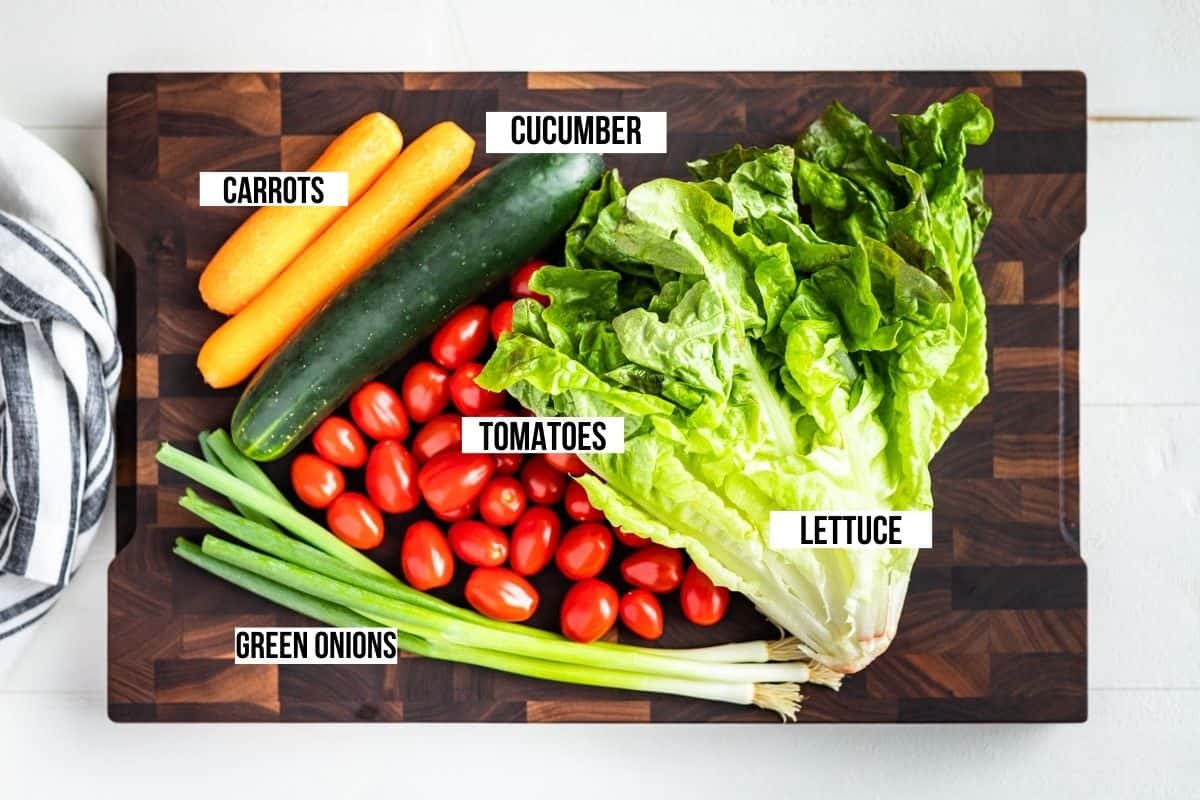 Green leaf lettuce, green onions, cherry tomatoes, cucumber, and carrots on a wooden cutting board.