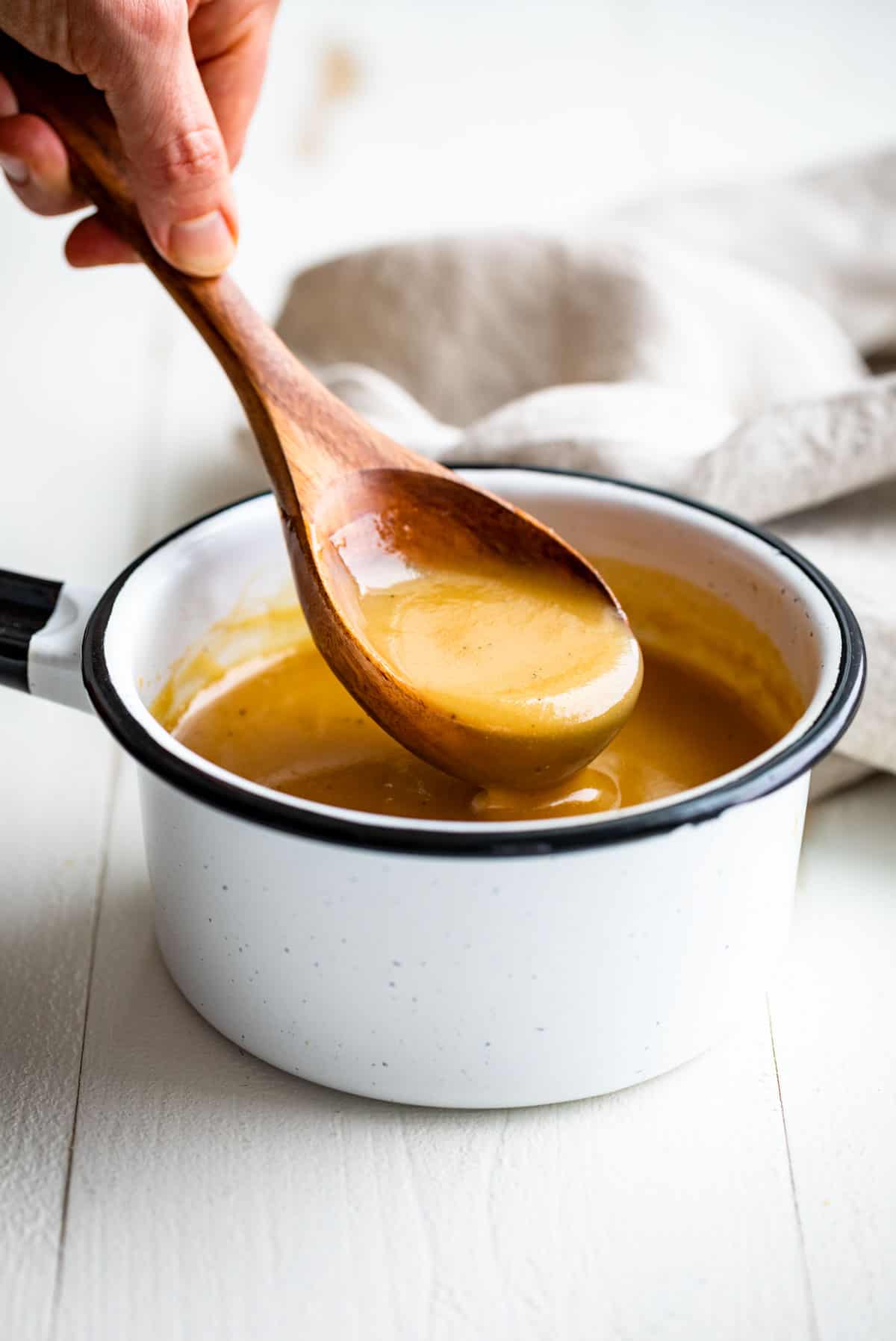 A hand holding a wooden spoon lifting out some finished gravy from a white pot.