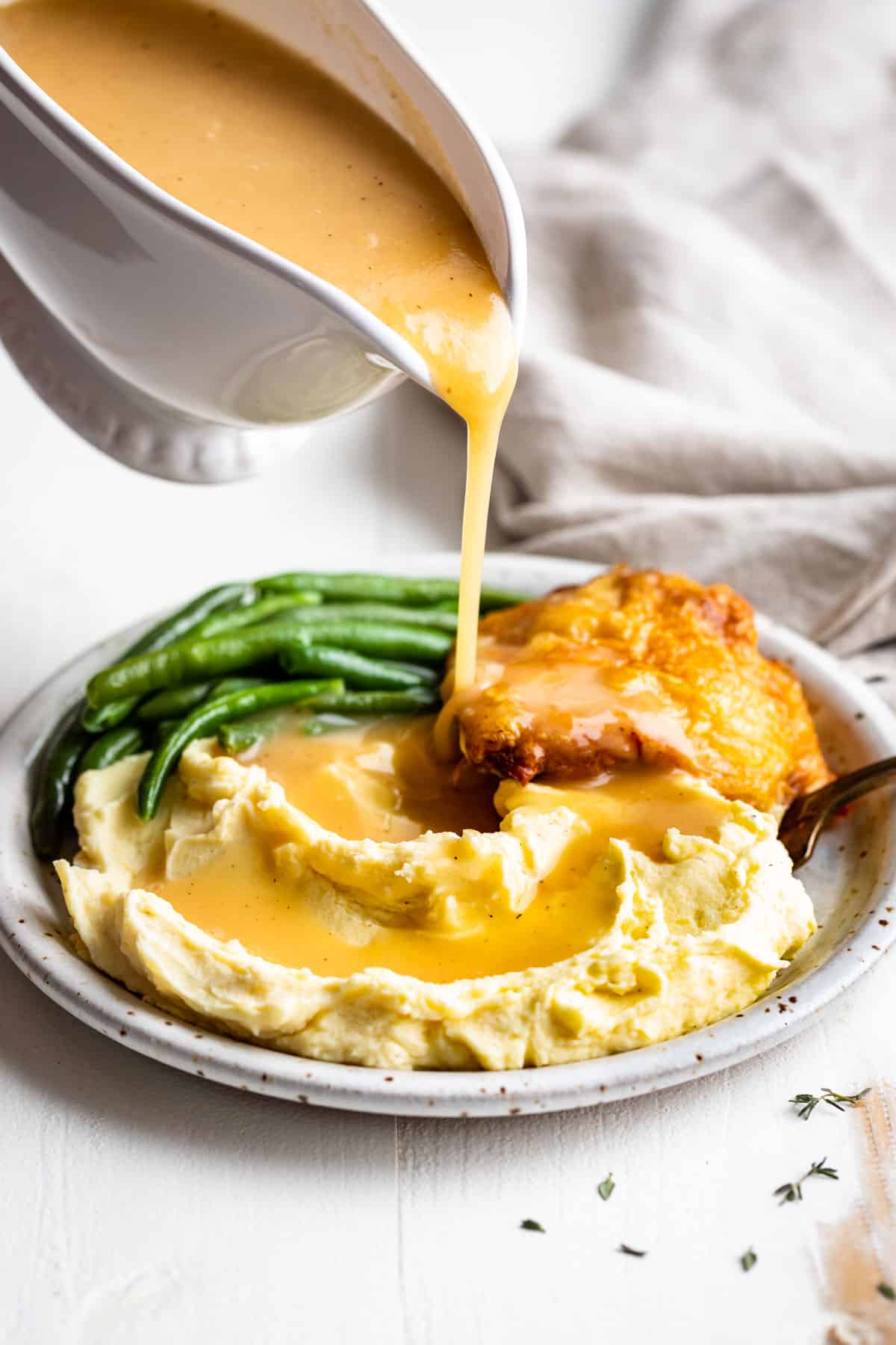 Gravy being poured out of a gravy boat onto mashed potatoes with green beans and chicken in the background.