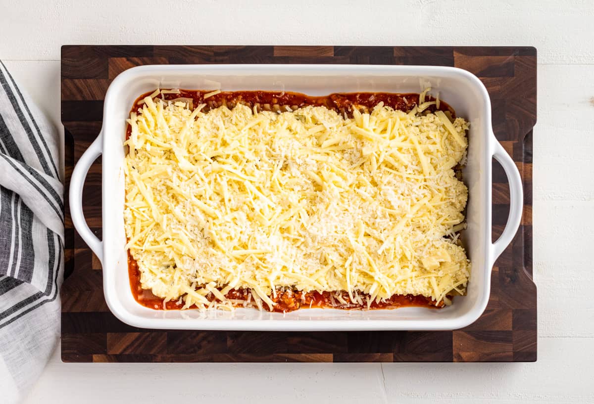 Ricotta and grated mozzarella layer over the sauce and lasagna noodles in a white baking dish.