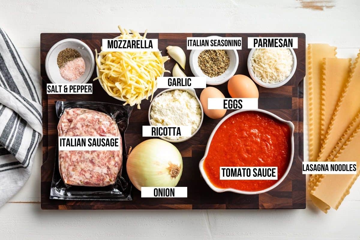 Italian sausage, onion, ricotta, mozzarella cheese, parmesan, tomato sauce, garlic, and lasagna noodles on a wooden cutting board.