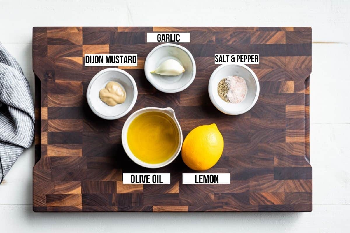 Olive oil, lemon, sea salt and pepper, garlic, and Dijon mustard on a wooden cutting board.