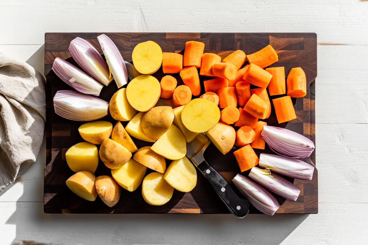 Cut up onion, potatoes, and carrots on a wooden cutting board.