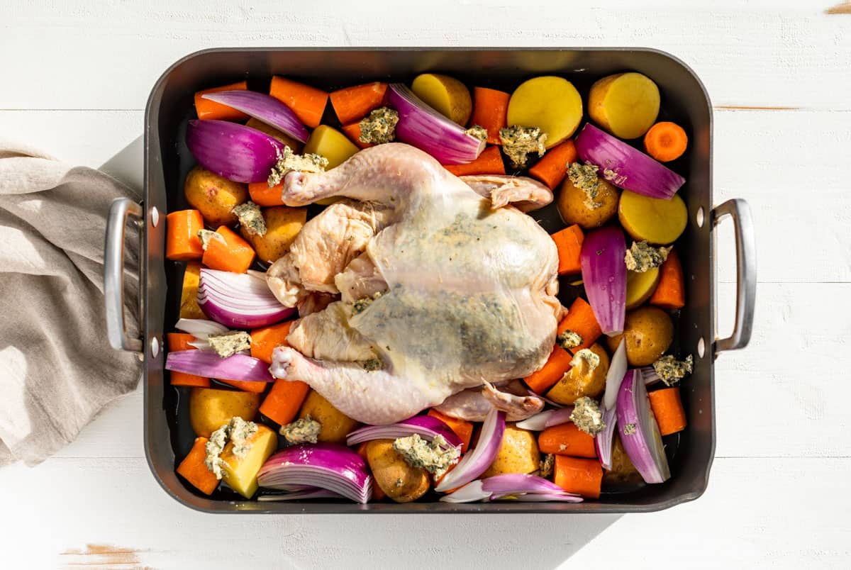 Cut up onion, carrots, and potatoes in a grey roaster with a whole chicken rubbed with herb butter in the center.