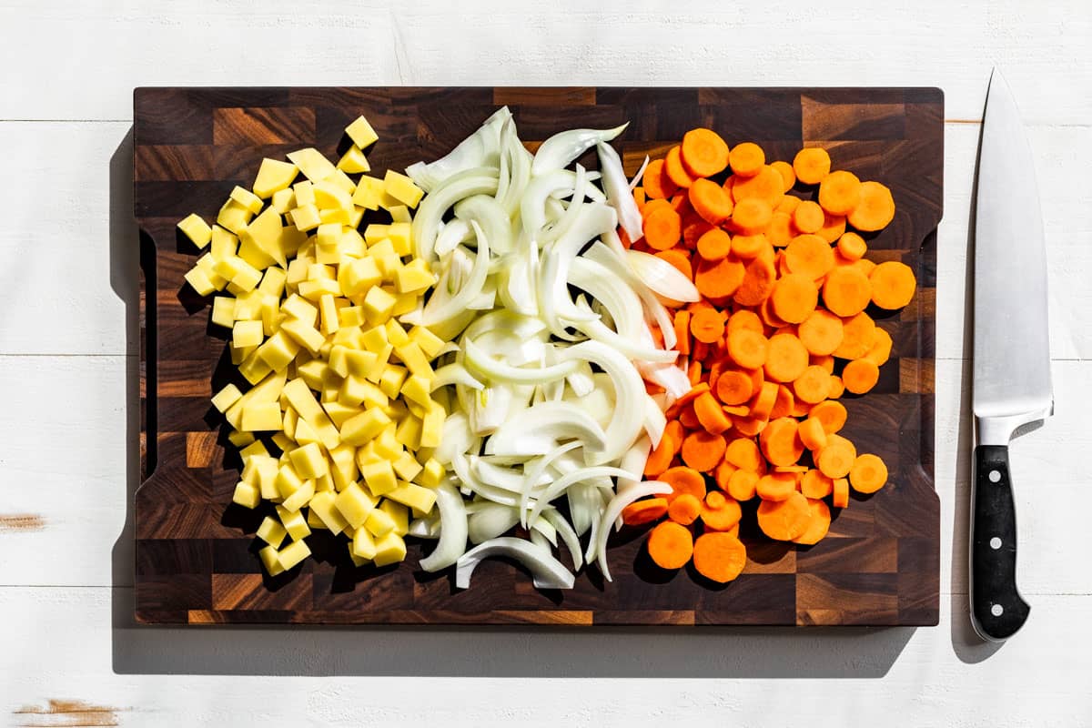 Diced potato, sliced onion, and slice carrots on a wooden cutting board.