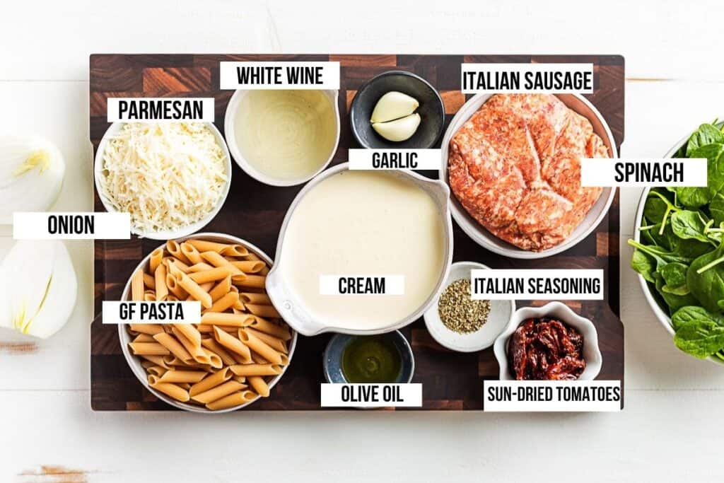 Gluten free pasta, parmesan, cream, white wine, onion, sundried tomatoes, Italian sausage, and spinach measured out in bowls on a cutting board.