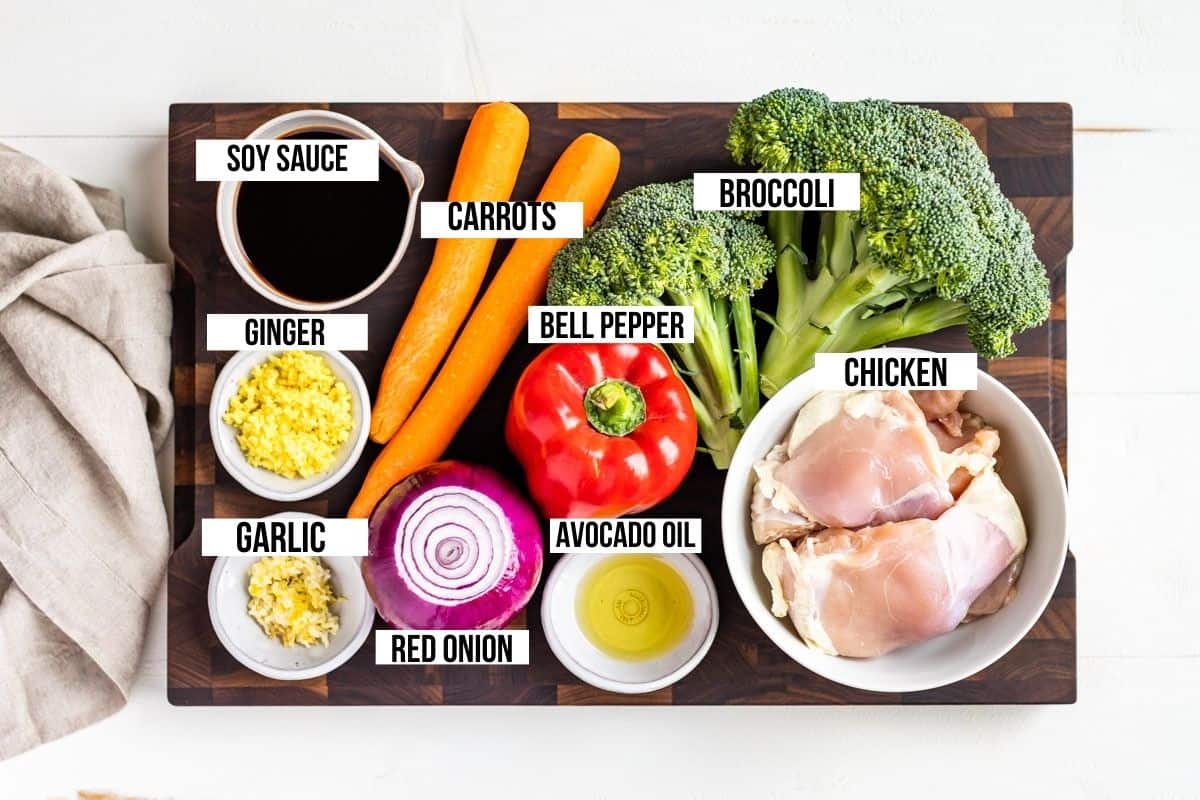 Broccoli, carrots, red onion, red bell pepper, ginger, garlic, coconut aminos, and chicken thighs in bowls on a wood cutting board.