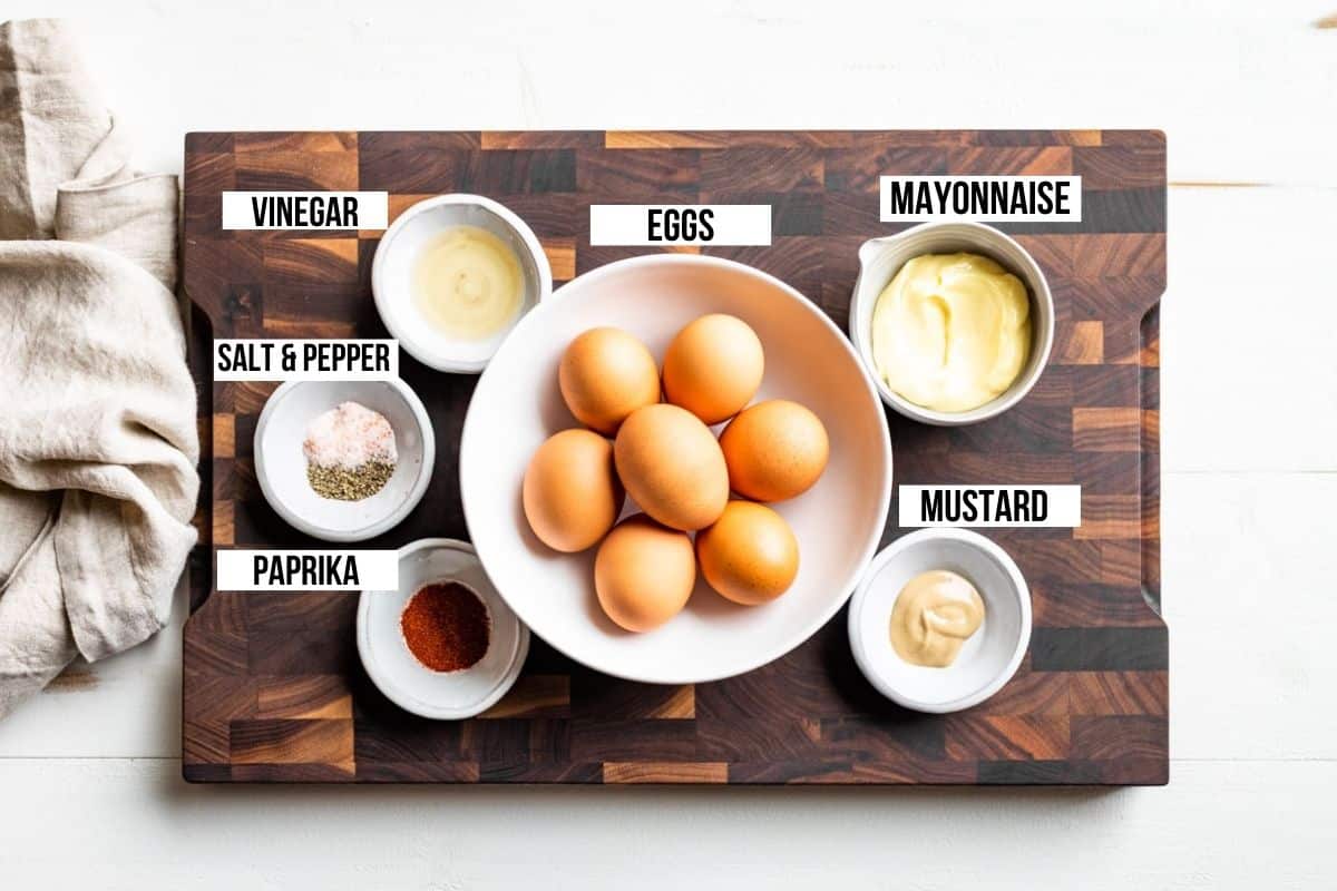 Eggs, mayonnaise, Dijon mustard, salt, pepper, paprika, and white wine vinegar on a wooden cutting board.