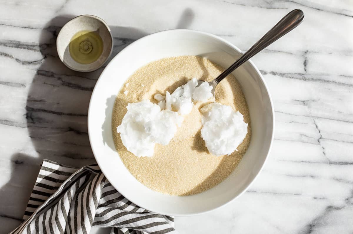 A large white bowl with cane sugar with the coconut oil being added with a silver spoon in it.